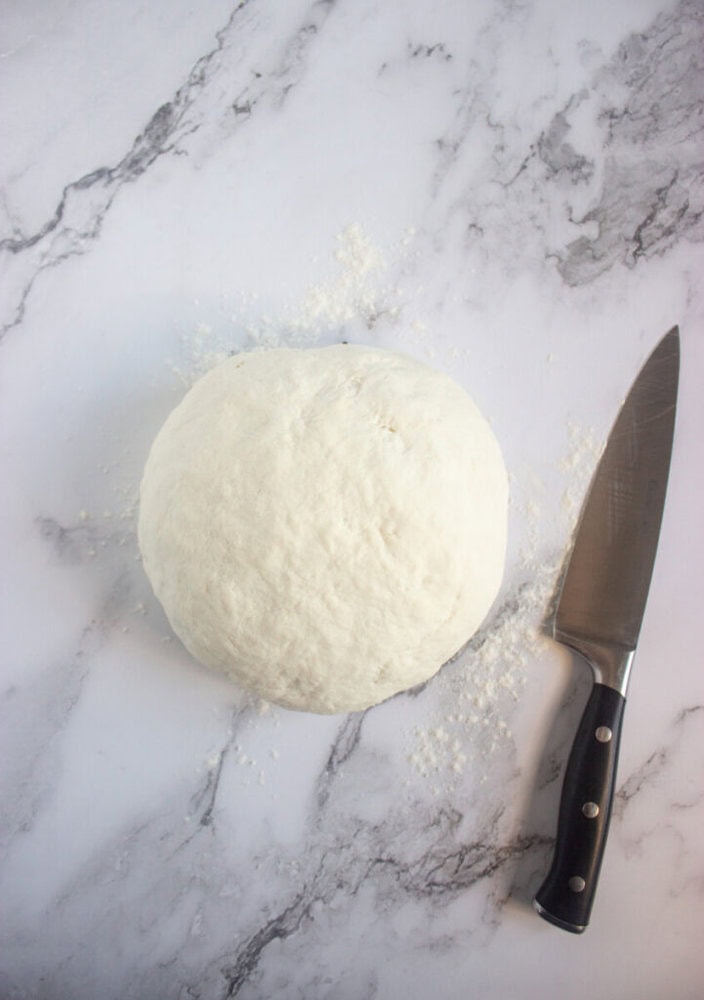 A ball of pizza dough sits on a floured marble surface next to a kitchen knife.