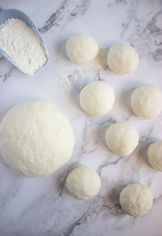 Balls of dough and a scoop of flour are arranged on a lightly floured marble surface.