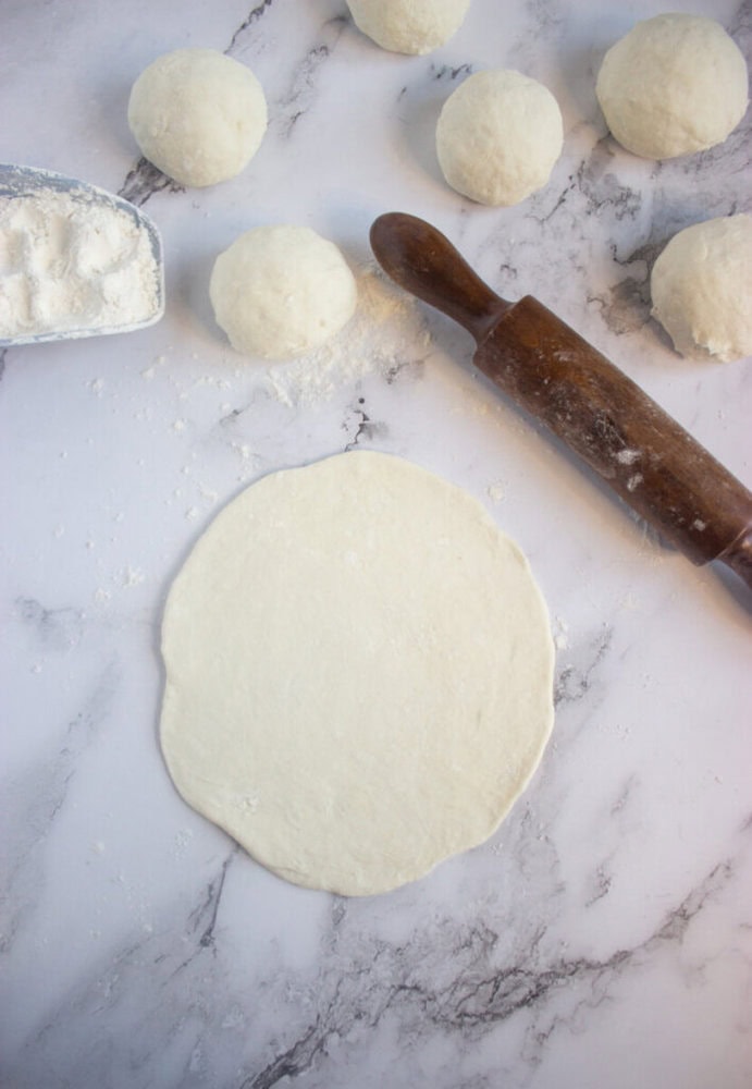 A flattened round piece of dough is on a floured surface, surrounded by dough balls, a rolling pin, and a metal scoop of flour.