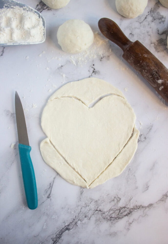 A sheet of dough cut into a heart shape sits on a marble surface beside a knife, rolling pin, flour, and dough balls.