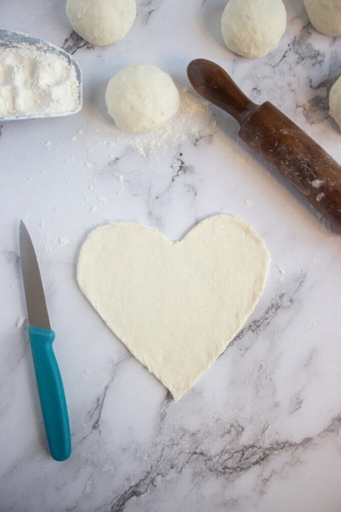 A heart-shaped piece of dough on a floured surface, with a rolling pin, a knife, dough balls, and a tray of flour nearby.