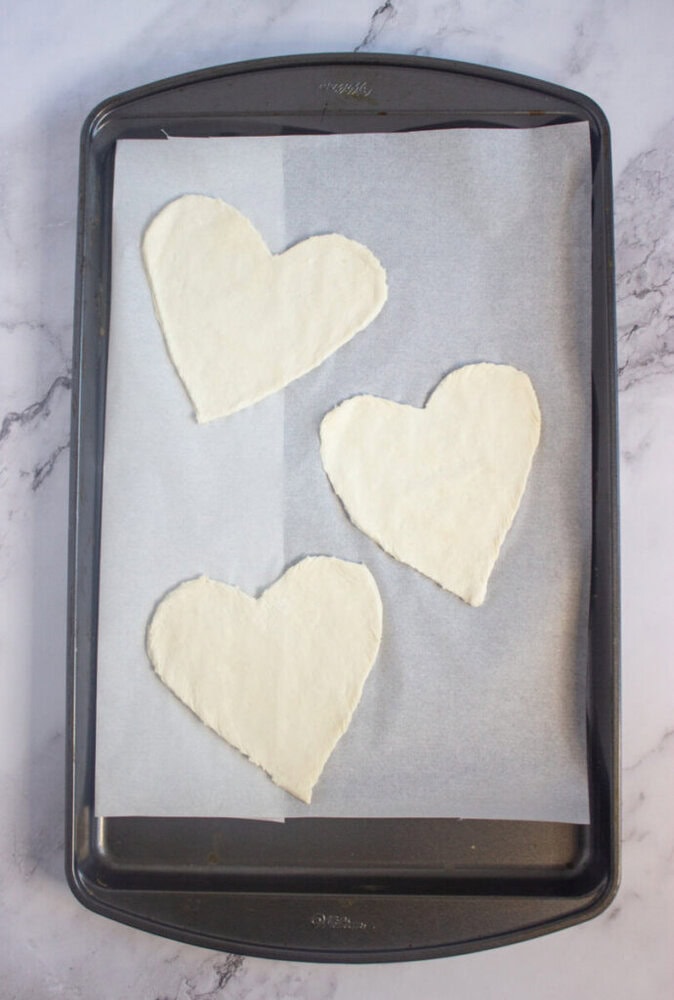 Three heart-shaped pieces of dough are placed on parchment paper on a baking sheet, ready to be baked.