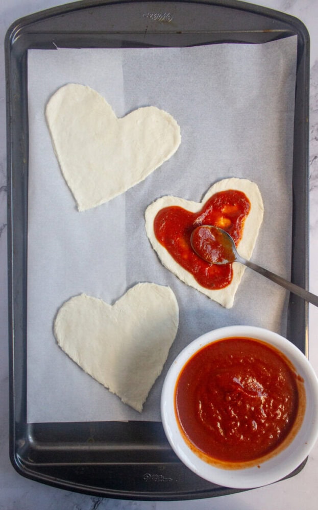 Baking tray with three heart-shaped pizza dough pieces, one being spread with tomato sauce, and a bowl of tomato sauce with a spoon.