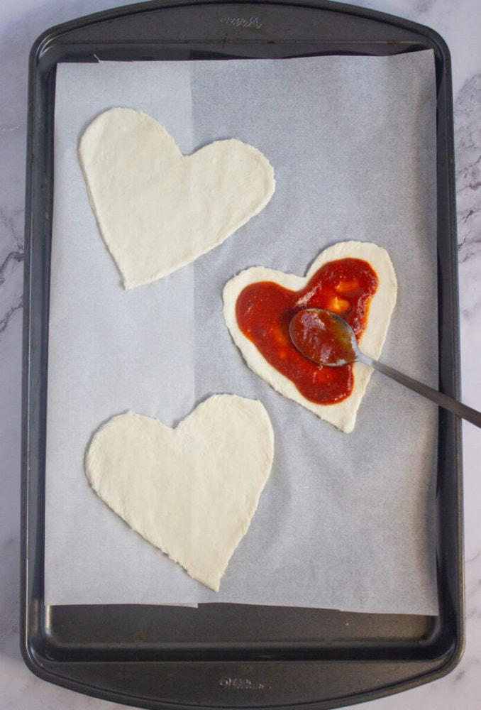A baking tray with three heart-shaped pieces of dough; one is being topped with tomato sauce using a spoon.