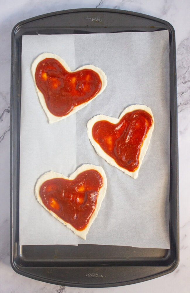 Three heart-shaped pizza doughs with tomato sauce on top are placed on a parchment-lined baking sheet.