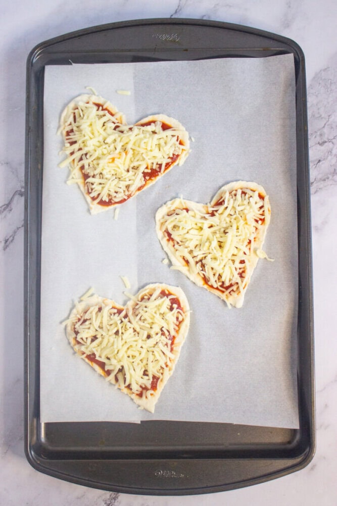 Three heart-shaped mini pizzas with tomato sauce and shredded cheese on parchment paper, placed on a baking tray.
