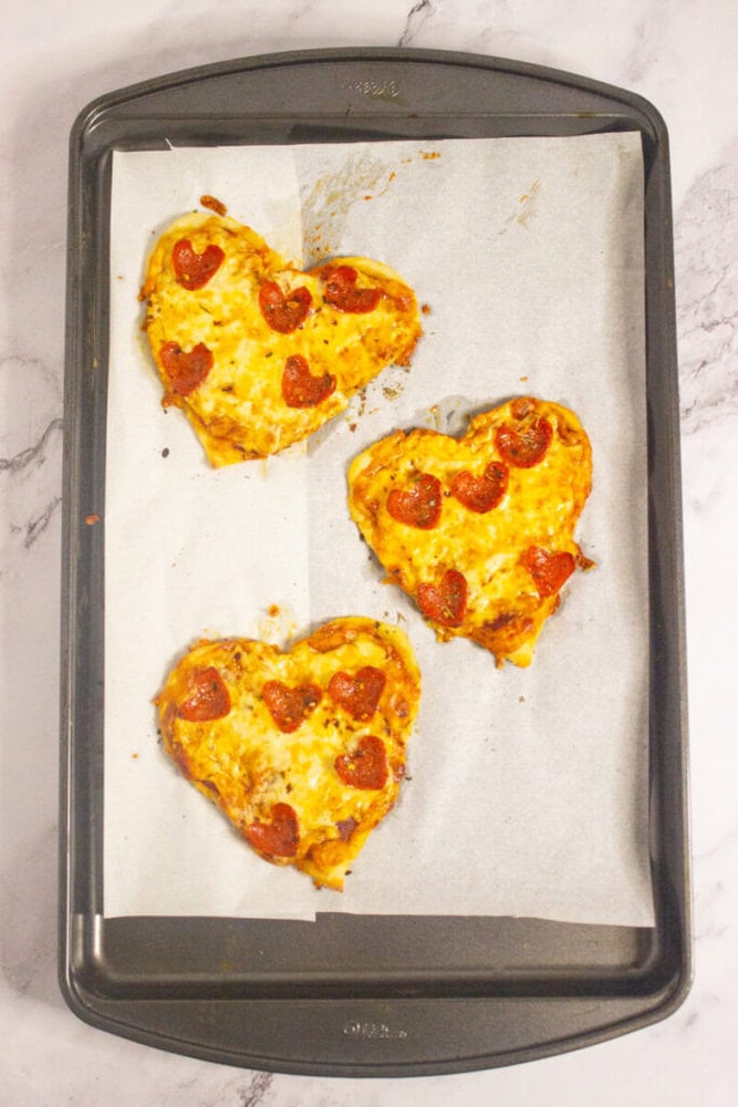 Three heart-shaped mini pizzas with pepperoni slices on parchment paper, placed on a baking sheet.
