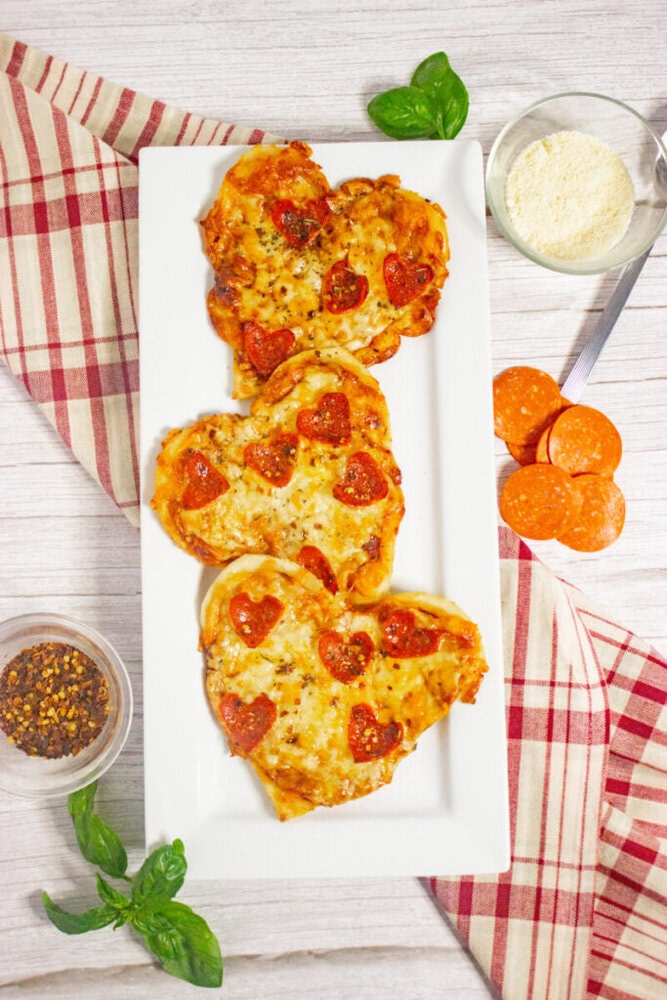 Three heart-shaped mini pizzas with heart-shaped pepperoni slices on a rectangular white plate, surrounded by basil, grated cheese, pepperoni, and seasonings on a striped cloth.