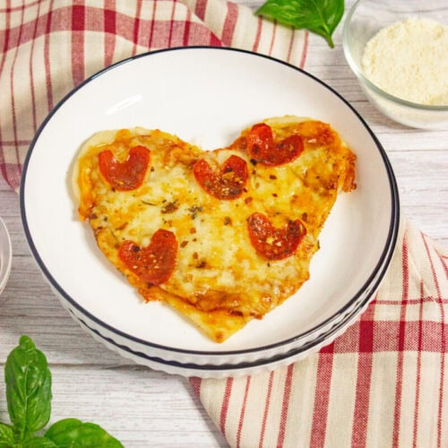 A heart-shaped pizza with pepperoni slices is served on a white plate with a striped rim, beside a red plaid cloth and fresh basil leaves.