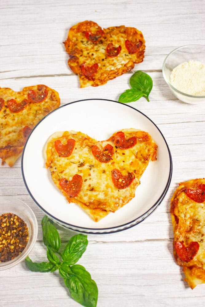 Heart-shaped pepperoni pizzas are arranged on a white wooden surface, with fresh basil leaves, grated cheese, and crushed red pepper flakes nearby. One pizza is on a white plate.