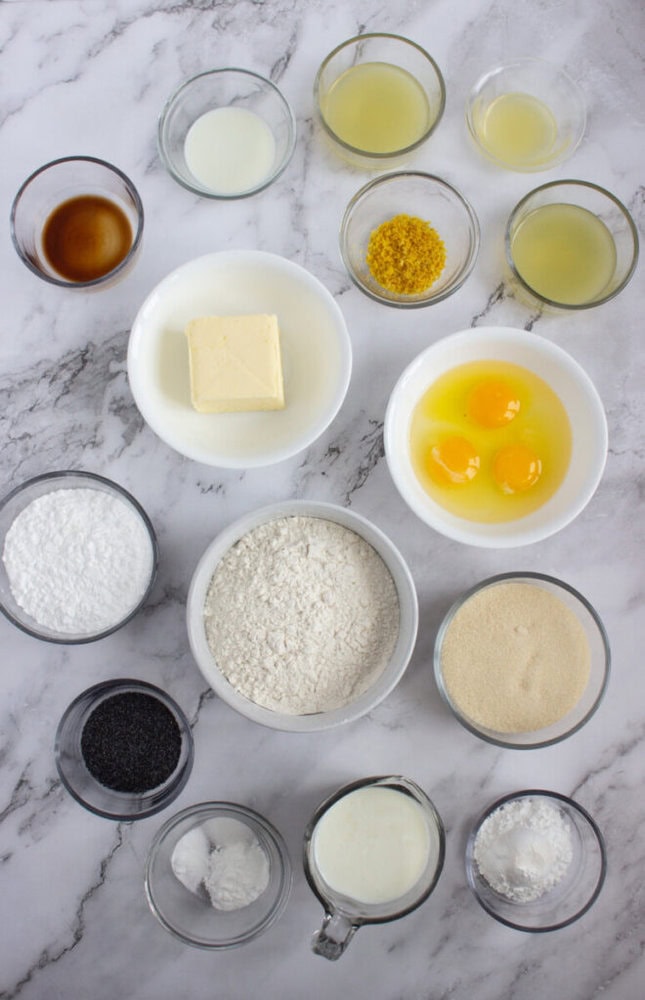 Various baking ingredients, including eggs, butter, flour, sugar, poppy seeds, lemon zest, and liquids, are arranged in small bowls on a marble countertop.