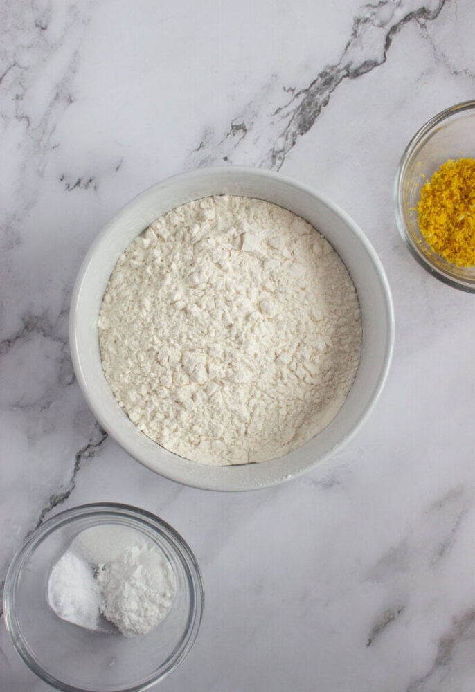 A bowl of flour is centered, surrounded by a small bowl of lemon zest and another small bowl containing salt and baking powder on a marble surface.