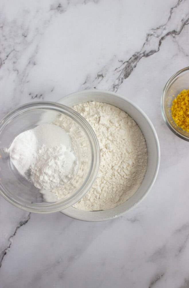 A bowl of flour and a glass bowl with baking powder and sugar on a marble surface, with a small bowl of lemon zest nearby.