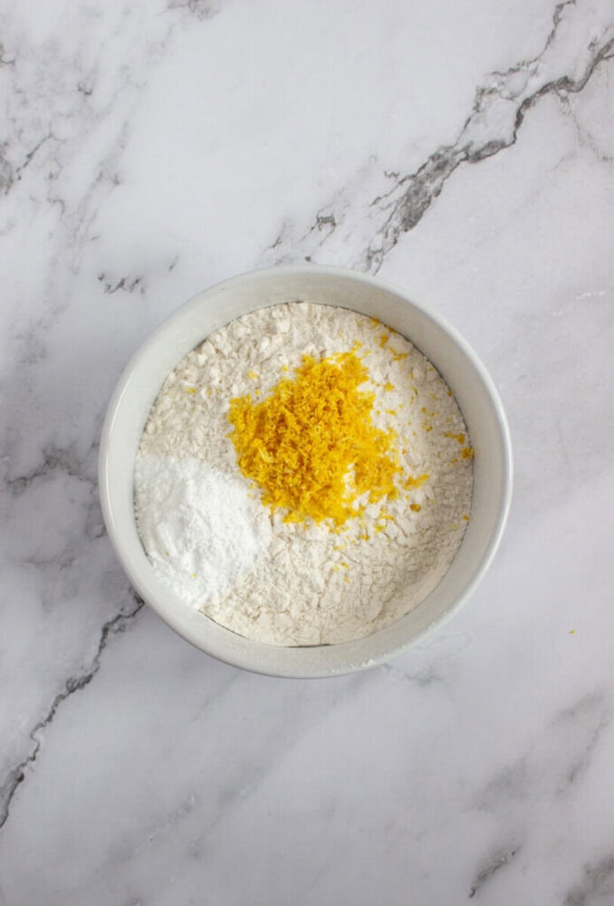 A white bowl containing flour, baking powder, and lemon zest sits on a white marble surface.