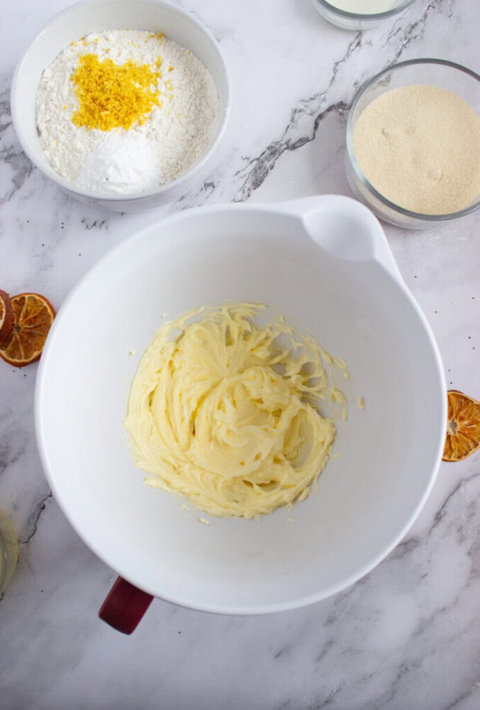 A white mixing bowl with creamed butter, surrounded by bowls of flour with lemon zest and granulated sugar on a marble countertop.