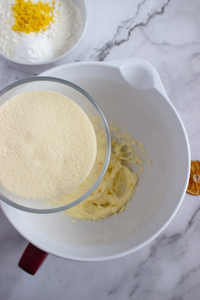 A bowl of softened butter is in a white mixing bowl. A glass bowl with semolina is being poured in. Another bowl with flour and lemon zest is nearby on a marble countertop.