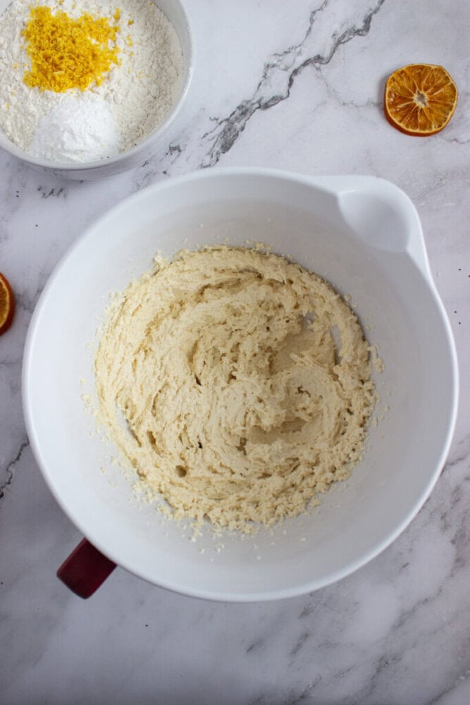 A white mixing bowl with creamed butter and sugar sits on a marble surface; nearby are a bowl of flour with lemon zest and slices of dried orange.