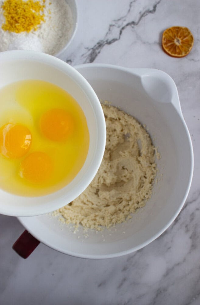 A white bowl with three raw eggs is held above another bowl containing creamed batter on a marble countertop. Flour and lemon zest are visible in a separate bowl, with a dried citrus slice nearby.