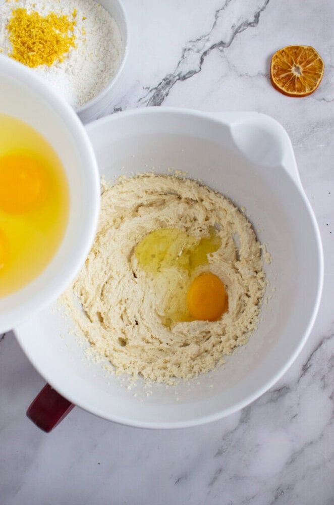 A mixing bowl with creamed butter and sugar, partially mixed with eggs. Another bowl with eggs is being poured in. Flour and orange zest are visible in a separate bowl.
