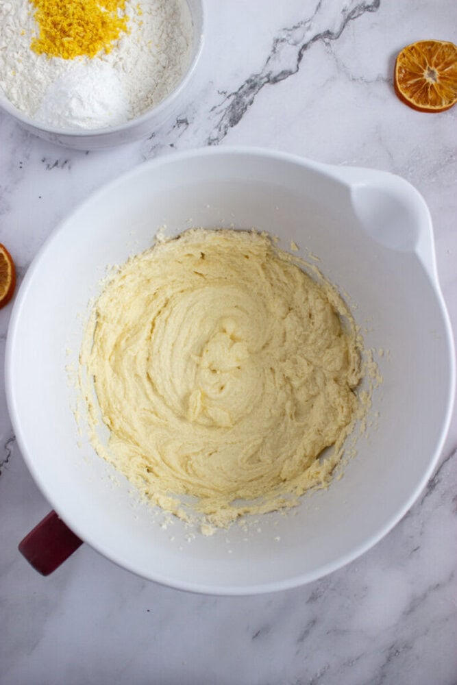 A white mixing bowl with creamed butter and sugar sits on a marble countertop, surrounded by a bowl of flour and dried orange slices.
