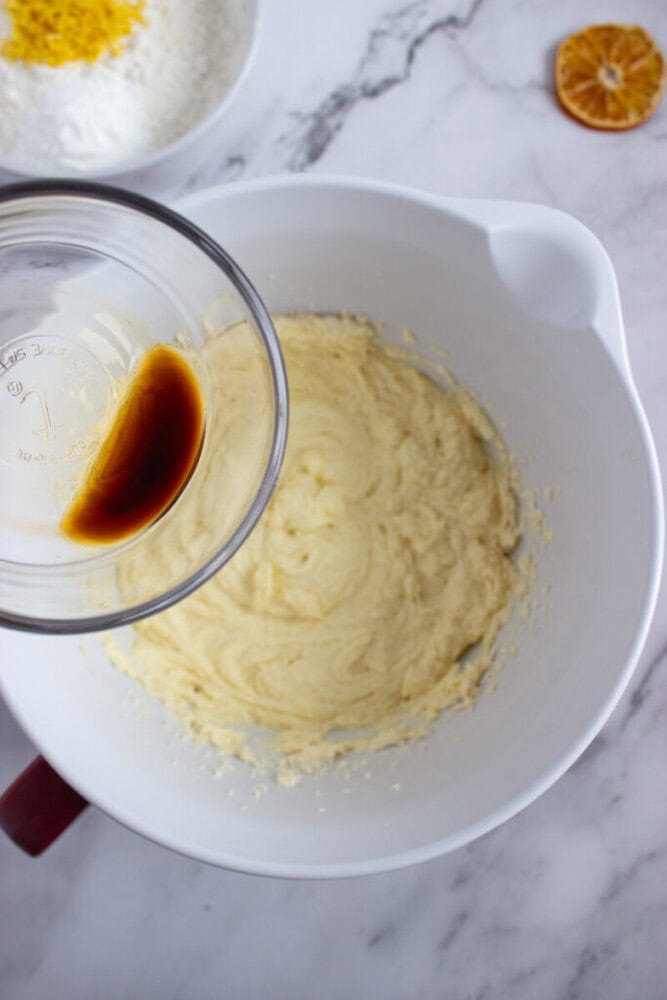 A glass cup with vanilla extract is about to be poured into a white mixing bowl containing creamed butter and sugar on a marble countertop.