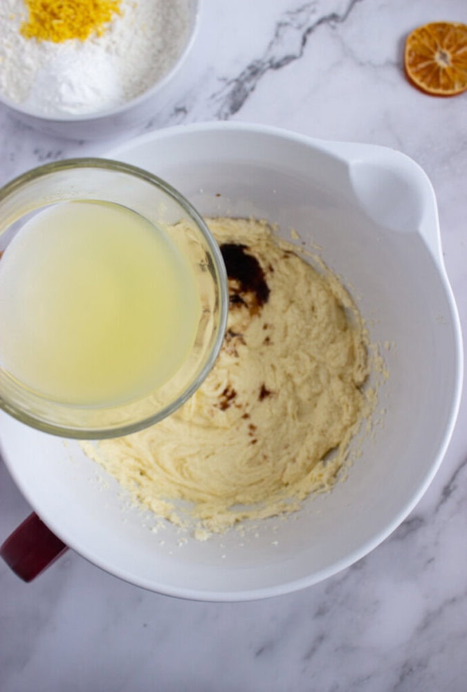 A glass bowl of lemon juice is being poured into a white mixing bowl containing creamed ingredients and vanilla extract on a marble countertop.