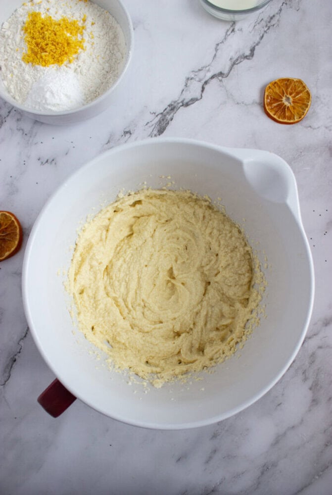 A mixing bowl with creamed batter sits on a marble surface next to a bowl of flour and orange zest, with dried orange slices nearby.
