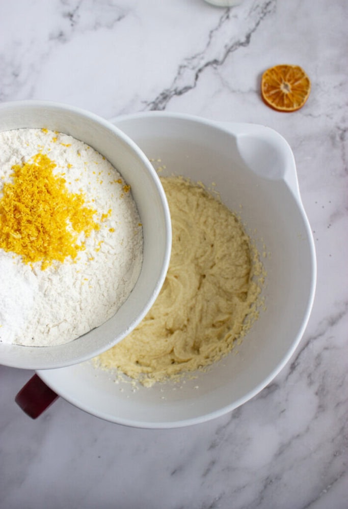 A bowl with flour and orange zest is being poured into a larger bowl containing a creamy batter on a marble countertop. A dried orange slice is visible nearby.