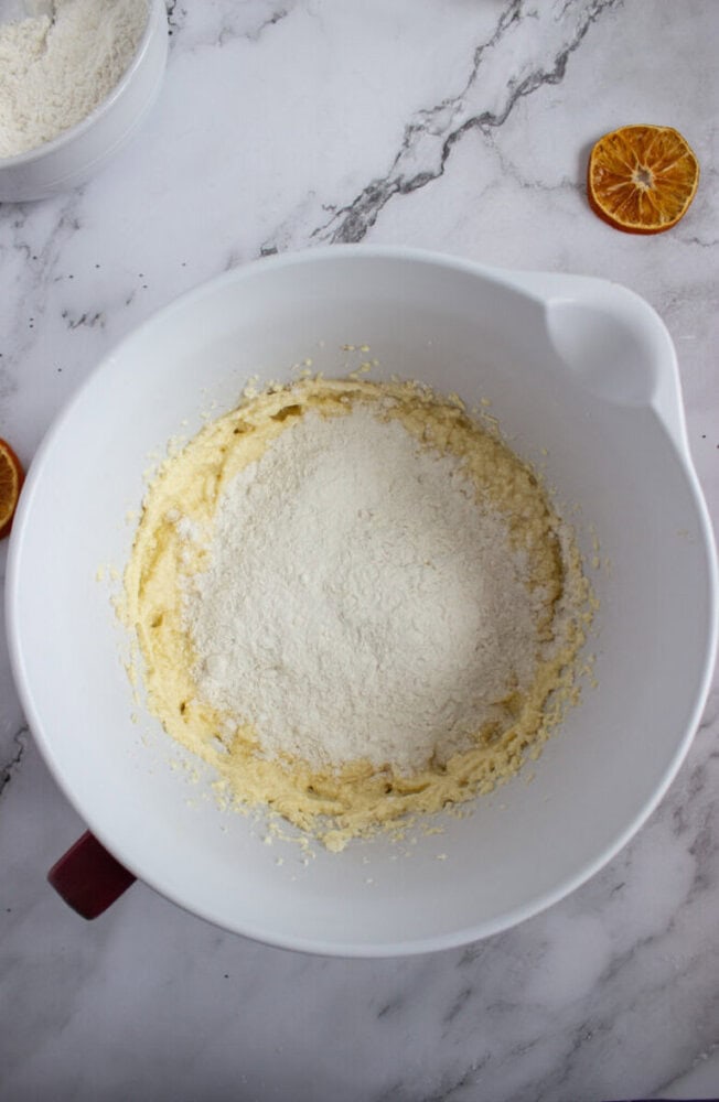 A white mixing bowl contains creamed batter with a mound of flour on top, set on a marble surface with dried orange slices nearby.