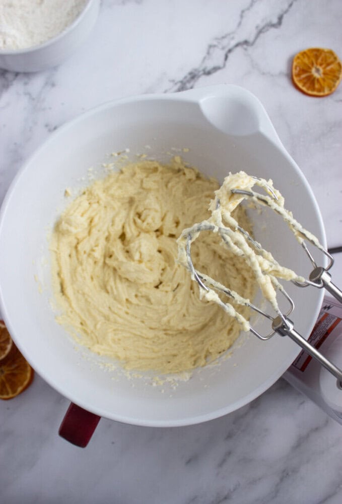 A white mixing bowl with creamed batter and an electric hand mixer with batter on the beaters, on a marble surface with dried orange slices nearby.