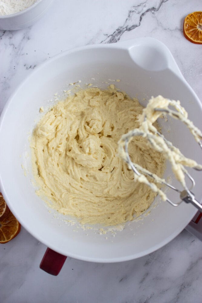 A white mixing bowl with creamed batter and a hand mixer, on a marble countertop with dried orange slices nearby.