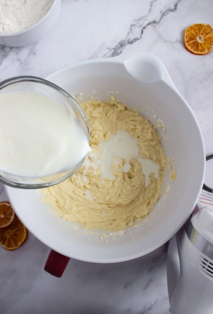 A bowl of batter is being mixed while milk is being poured in from a glass container; a mixer, flour bowl, and dried orange slices are nearby.