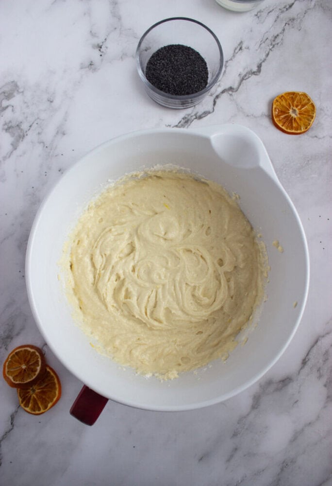 A white mixing bowl with cake batter sits on a marble surface, surrounded by dried orange slices and a small glass bowl of poppy seeds.