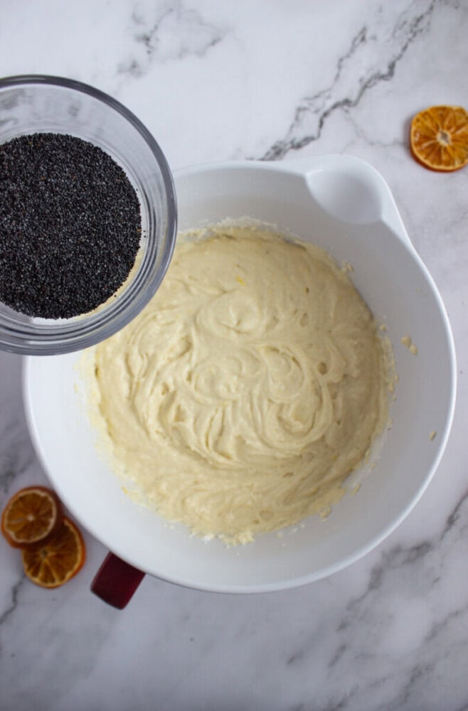 A bowl of cake batter sits on a marble surface next to dried orange slices; a hand holds a cup of poppy seeds above the batter, ready to add them.