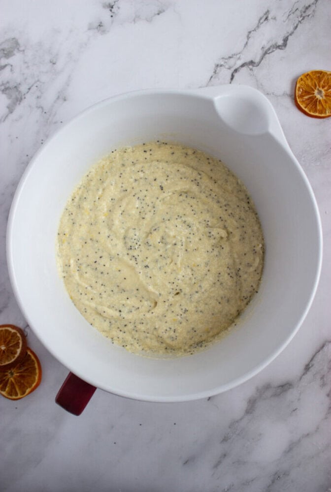 A white mixing bowl with poppy seed cake batter sits on a marble surface, surrounded by a few slices of dried orange.