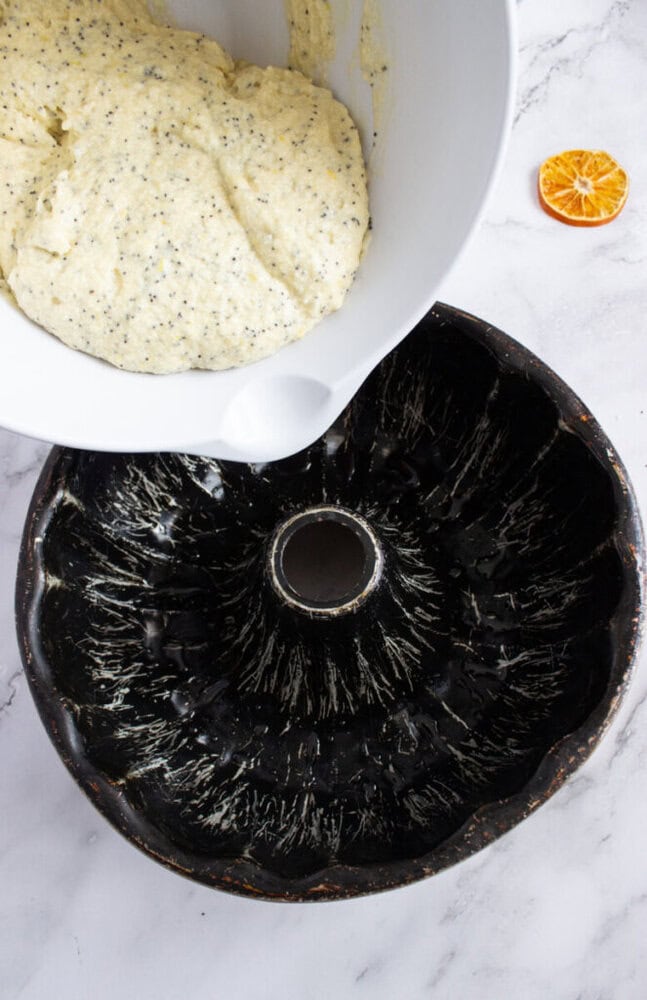 A white mixing bowl with cake batter is being poured into a greased bundt pan; a sliced orange sits on the marble countertop nearby.