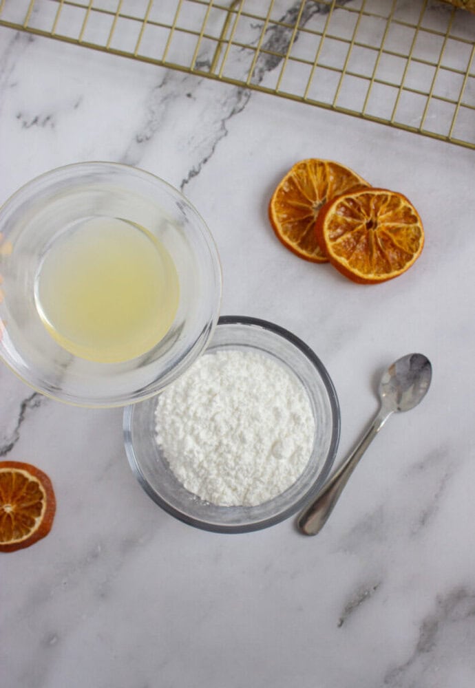 A bowl of powdered sugar, a spoon, dried orange slices, and a glass bowl containing lemon juice are arranged on a marble surface near a cooling rack.