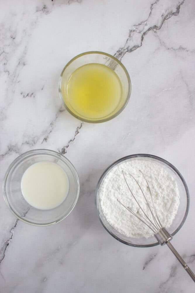 Three bowls on a marble surface: one with yellow liquid, one with white liquid, and one with powdered sugar and a whisk.