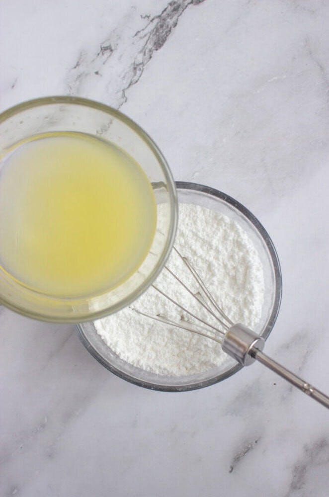A bowl of powdered sugar with a whisk next to a glass bowl containing lemon juice on a marble surface.