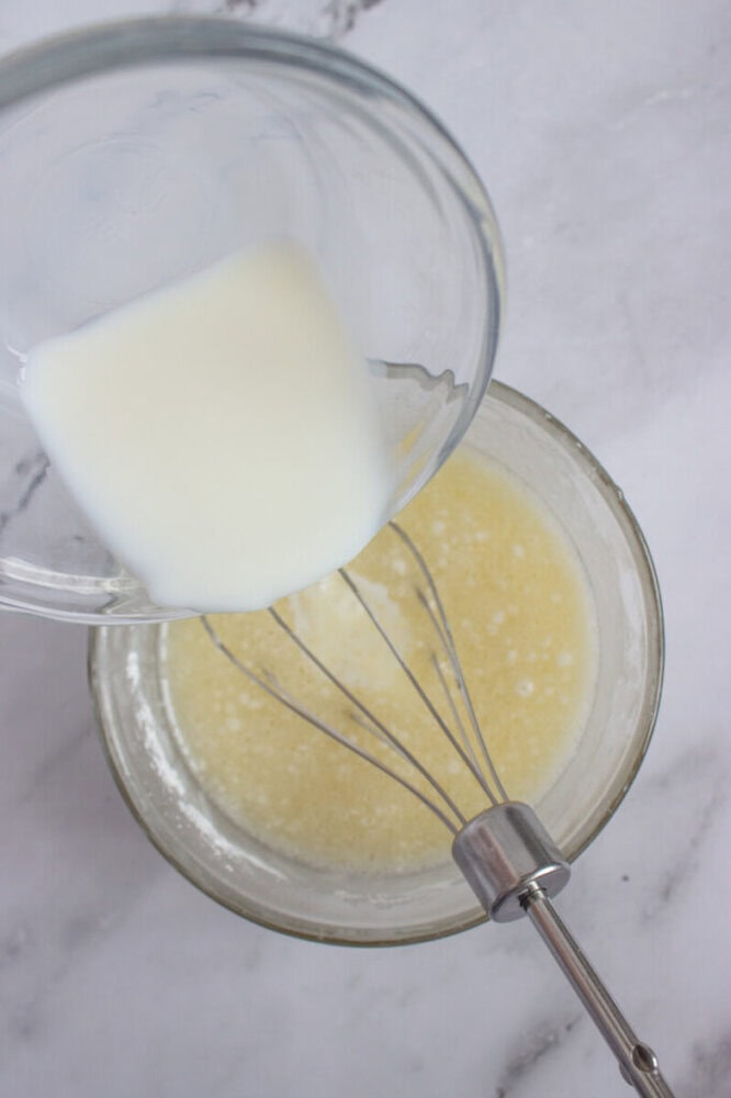 A glass bowl with melted butter is being mixed with milk from a measuring cup using a whisk on a marble surface.