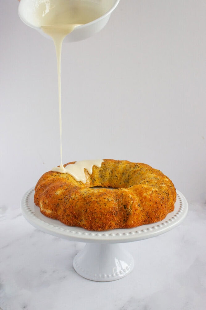A poppy seed bundt cake on a white cake stand being drizzled with white icing from a bowl above.