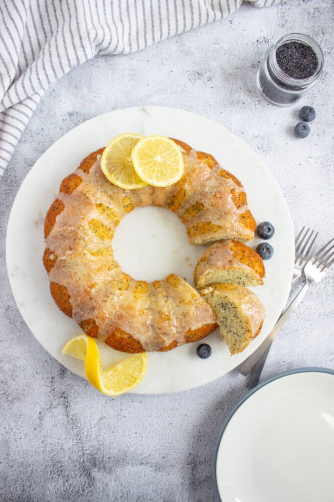 A lemon poppy seed bundt cake with glaze, garnished with lemon slices and blueberries, is displayed on a white marble plate next to forks, a napkin, and an empty plate.