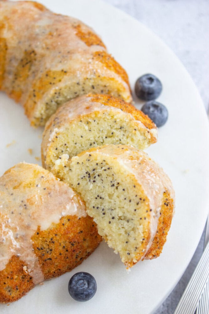 A sliced poppy seed bundt cake with a glaze on top is displayed on a white surface with a few blueberries beside it.