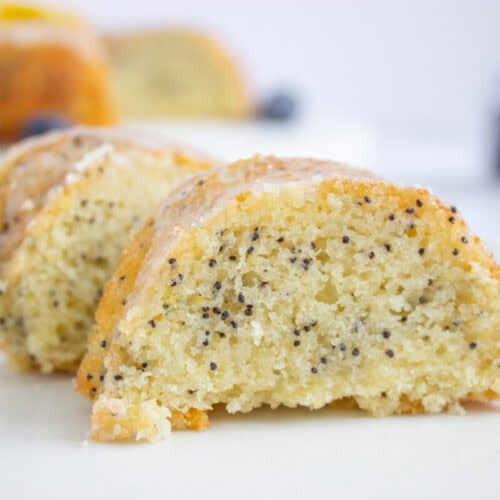 Two slices of poppy seed cake with a light glaze are displayed on a white plate, showing a moist, crumbly texture and visible poppy seeds.