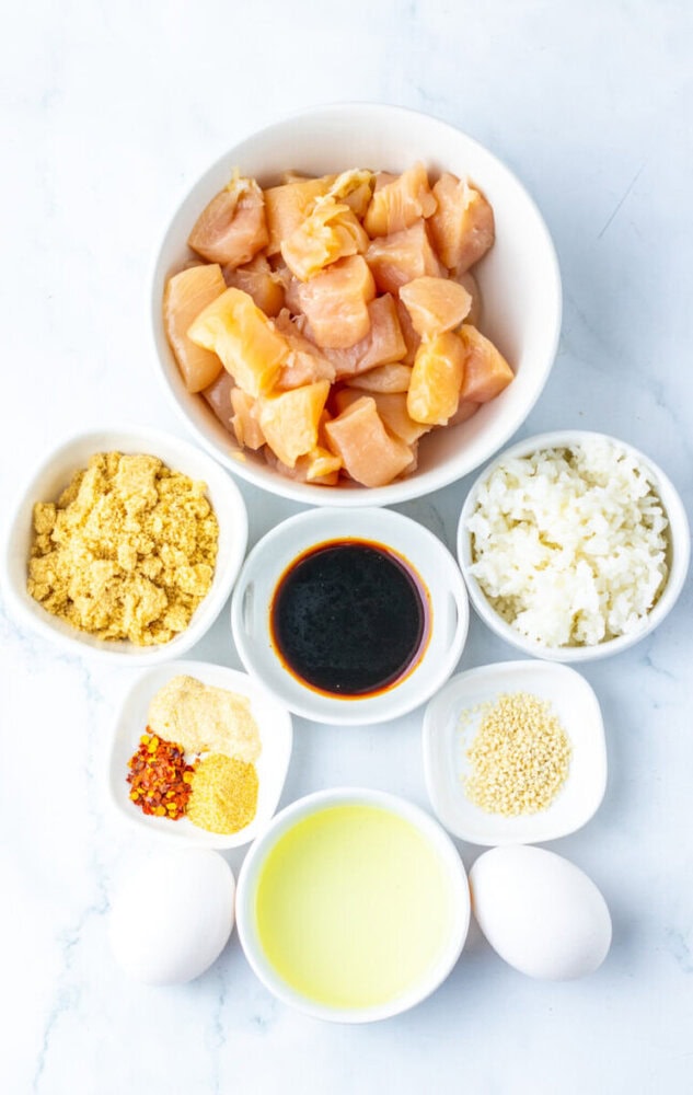 Top-down view of bowls containing raw diced chicken, cooked rice, soy sauce, cornstarch, spices, sesame seeds, beaten eggs, and two whole eggs on a white surface.