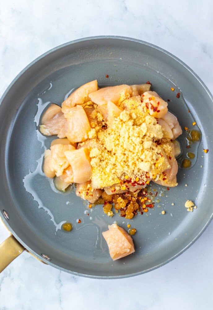 Chunks of raw chicken breast in a frying pan topped with dry seasoning and chili flakes, ready to be cooked.