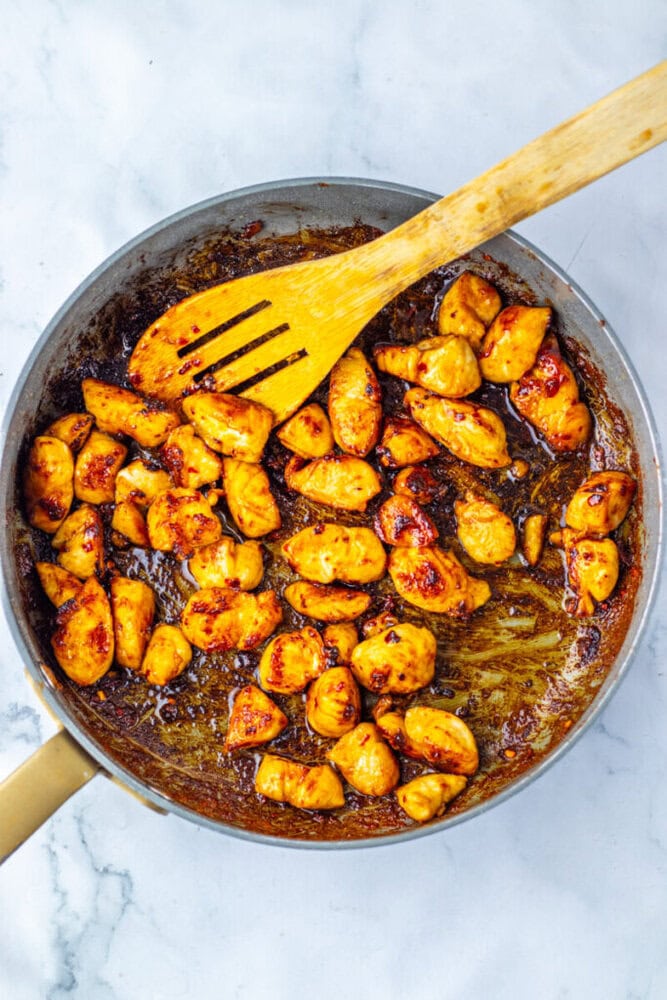 Cooked, browned chicken pieces in a skillet with sauce, stirred by a wooden spatula on a marble countertop.