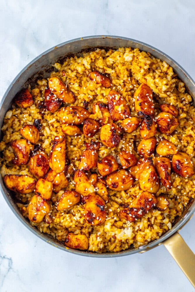 A skillet filled with yellow fried rice topped with glazed chicken pieces and sprinkled with sesame seeds, on a white marble surface.