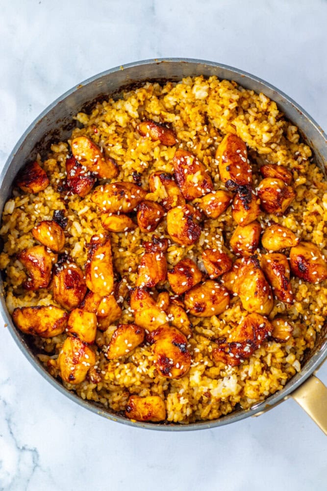 A pan filled with golden fried rice and pieces of glazed chicken, garnished with sesame seeds, on a marble surface.