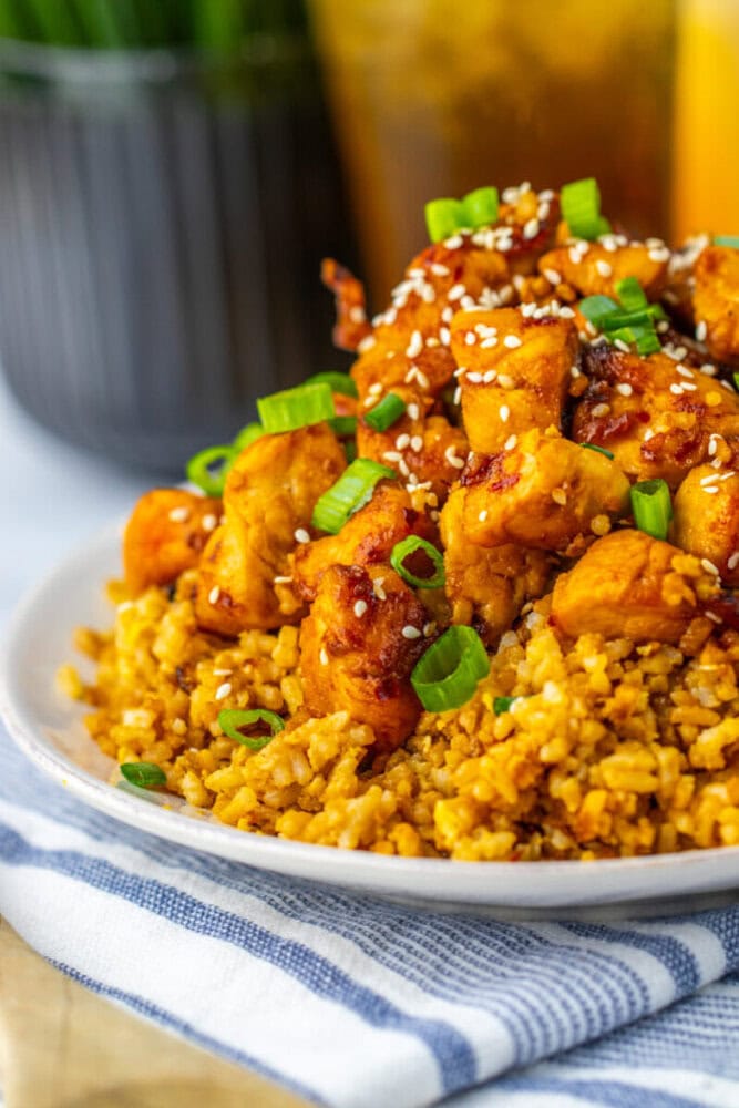 A plate of seasoned rice topped with pieces of glazed chicken, garnished with sesame seeds and chopped green onions.